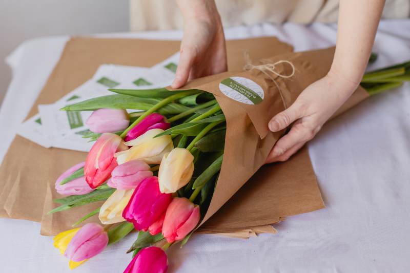 Gruppo di capre domestiche bianche e marroni che guardano verso la fotocamera in un prato verde della fattoria, con alberi e strutture agricole sullo sfondo sotto un cielo nuvoloso