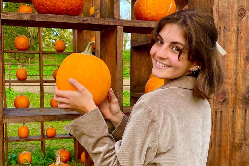 Giovane donna sorridente che tiene una zucca arancione davanti a una struttura espositiva in legno piena di zucche di varie dimensioni in un ambiente naturale autunnale