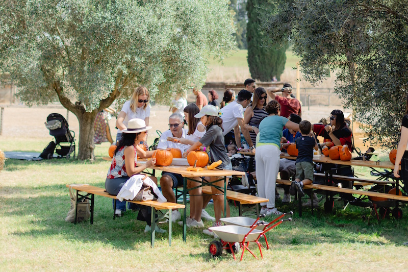 Famiglie sedute ai tavoli di legno all'aperto sotto gli alberi durante i laboratori di lavorazione delle zucche, con bambini e adulti che partecipano alle attività creative in un ambiente naturale
