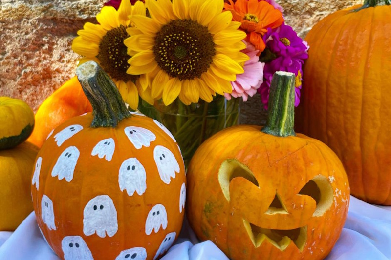 Elegante allestimento autunnale con tavolo di ferro battuto bianco decorato con ceste di vimini piene di zucche arancioni, lanterne bianche, mazzi di grano e foglie colorate, con persone sullo sfondo che godono dell'atmosfera dell'evento