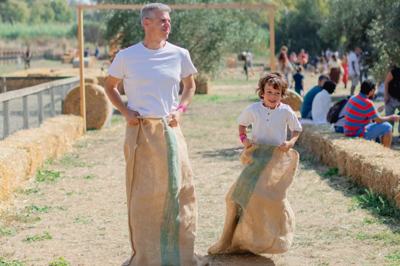 Papà e bambino che partecipano insieme a una gara con i sacchi di juta su un prato, con altri visitatori e balle di fieno sullo sfondo durante l'evento autunnale