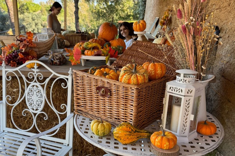 Elegante allestimento autunnale con tavolo di ferro battuto bianco decorato con ceste di vimini piene di zucche arancioni, lanterne bianche, mazzi di grano e foglie colorate, con persone sullo sfondo che godono dell'atmosfera dell'evento